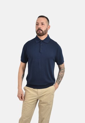 Uomo con tatuaggi che indossa una polo blu navy a maniche corte e pantaloni beige, in piedi davanti a uno sfondo chiaro e semplice.