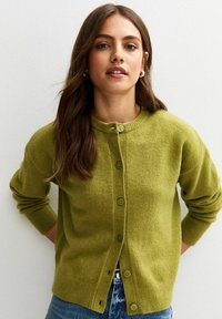 Groene cardigan van zacht materiaal, met een ronde halslijn, een knoopsluiting aan de voorkant en geribde manchetten. De textuur lijkt gezellig en warm.