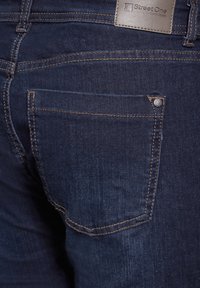Jean en denim bleu foncé avec une poche arrière présentant des coutures contrastantes et un détail de bouton argenté. Étiquette visible sur la taille.