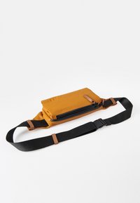 Gelbe Tasche mit einem Reißverschlussfach und einem verstellbaren schwarzen Tragegurt. Glatte Textur, rechteckige Form und Akzente aus braunem Leder.