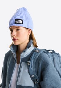 Gorro de punto acanalado color azul claro con un parche de logo negro, llevado con una chaqueta borrosa azul marino con un panel azul claro y correas de mochila.