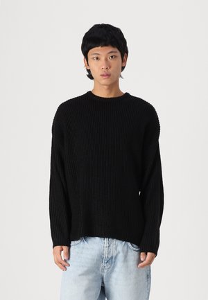 Black knitted sweater featuring a ribbed design, crew neckline, and loose fit, paired with light blue denim jeans.