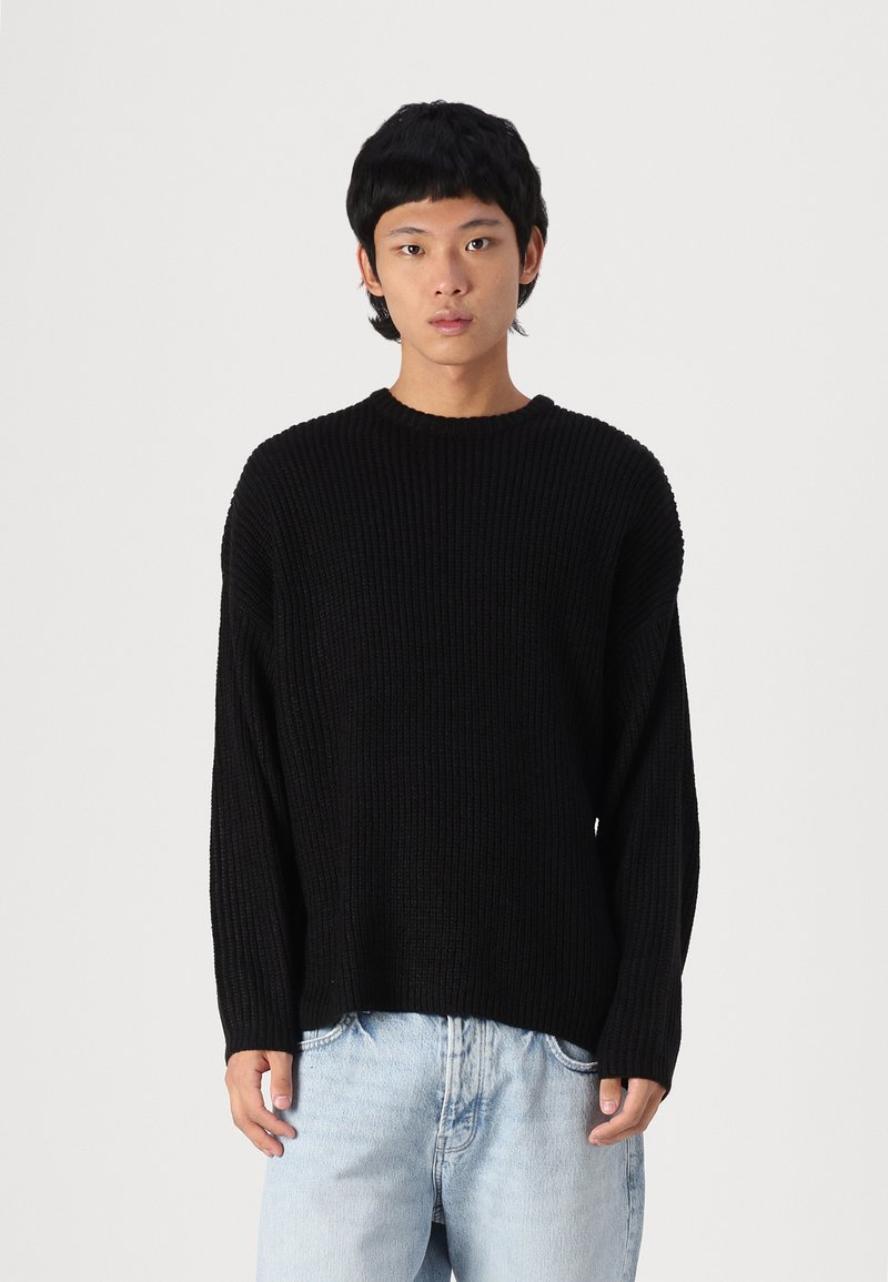Suéter negro de punto con diseño de relieve, cuello redondo y corte holgado, combinado con jeans de mezclilla azul claro.