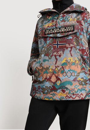 Pull Napapijri multicolore à motifs avec poche avant et patch du drapeau norvégien, porté avec un pantalon noir et les mains dans les poches.
