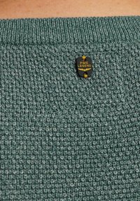 Pull en tricot vert foncé avec une surface texturée. Dispose d'une étiquette noire avec "PME LEGEND" en lettres dorées, fixée au niveau du col.
