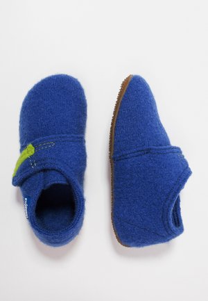 Paire de chaussons en laine bleue avec détails de couture verts et semelles en caoutchouc marron, vus de dessus et de côté sur fond blanc.