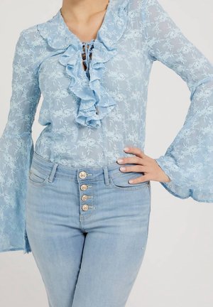 Femme portant un chemisier transparent bleu clair à motif floral avec des volants et des manches évasées, associé à un jean taille haute bleu clair avec plusieurs boutons.