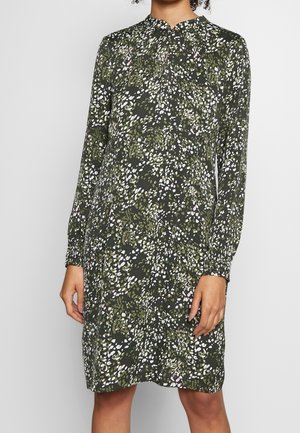 Kvinde iført knælængde, langærmet mørkegrøn kjole med hvidt og lyserødt abstrakt blomsterprint, stående foran ensfarvet baggrund.