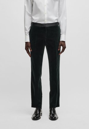 Pantalons en velours vert foncé avec une coupe ajustée, présentant une texture lisse et une taille noire, associés à des chaussures de ville noires.