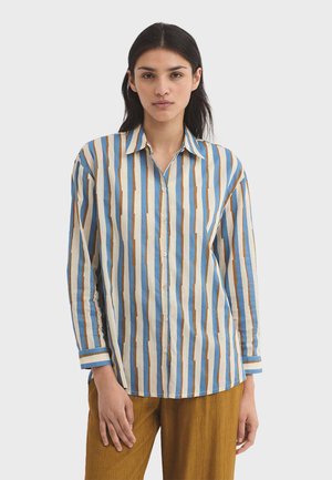 Frau trägt ein langärmeliges Hemd mit vertikalen blauen, weißen und braunen Streifen, kombiniert mit senffarbenen Hosen.