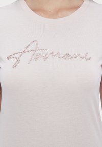 T-shirt rosa chiaro in cotone, con un design in rilievo di perline che scrive "Armani" sopra "EXCHANGE" in un font sottile.