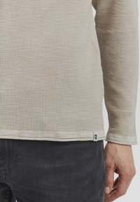 Beige long-sleeve shirt with a textured fabric and a subtle pattern, featuring a black logo tag at the hem. Paired with dark jeans.