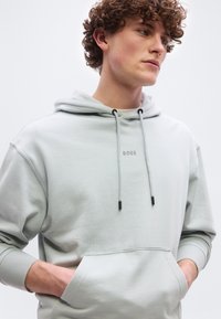 Νεαρός με κατσαρά μαλλιά που φοράει ανοιχτό γκρι φούτερ με το λογότυπο "BOSS", τα χέρια στις μπροστινές τσέπες, κοιτάζει δεξιά σε απλό φόντο.