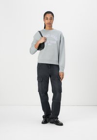 Felpa grigia con testo viola, abbinata a pantaloni cargo scuri con tasche laterali. Una borsa nera e delle scarpe completano il look.