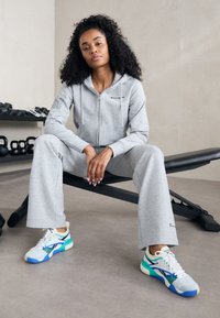 Hanorac gri cu fermoar și pantaloni asortați, confecționați dintr-un material moale. Încălțăminte sport colorată cu accente albastre, verzi și albe. Setare de sală de sport în interior.