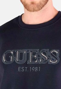 Hombre con camiseta negra de cuello redondo con un gran logo bordado de "GUESS" y el texto "EST. 1981" en el pecho.