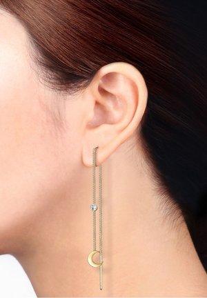 Gros plan sur l'oreille d'une femme portant une boucle d'oreille en chaîne dorée avec une petite pierre précieuse et un charm en forme de croissant de lune.