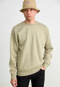Olivengrøn sweatshirt med rund hals og lange ærmer, med et hvidt Jordan-logo på brystet. Sorte bukser, neutral baggrund.