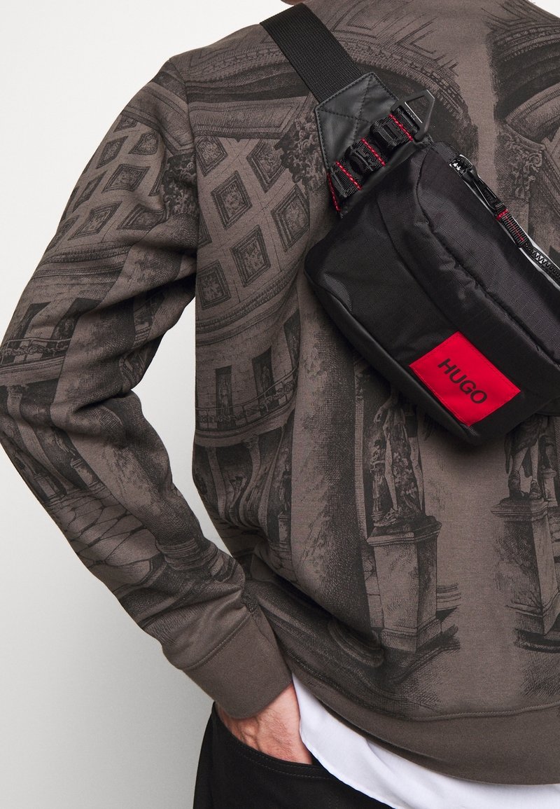 Sac à bandoulière noir avec logo "HUGO" rouge, présentant une finition texturée, une sangle réglable et une fermeture éclair, associé à un sweat-shirt marron à motifs.