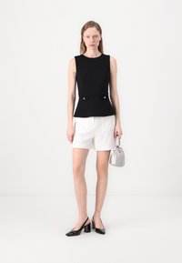 Top negru fără mâneci cu design peplum și două butoane în față, pantaloni scurți albăstrelor croiți, tocuri negre din patent și o geantă mică metalică.