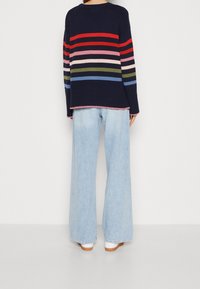 Marineblauwe trui met strepen in rood, roze, groen en blauw, gecombineerd met lichtblauwe jeans met brede pijpen en bruine schoenen.