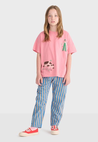 Młoda osoba ubrana w różowy t-shirt z ilustracjami rolnika i krowy, niebiesko-pomarańczowe paskowane spodnie oraz czerwone wsuwane buty z stokrotkami.