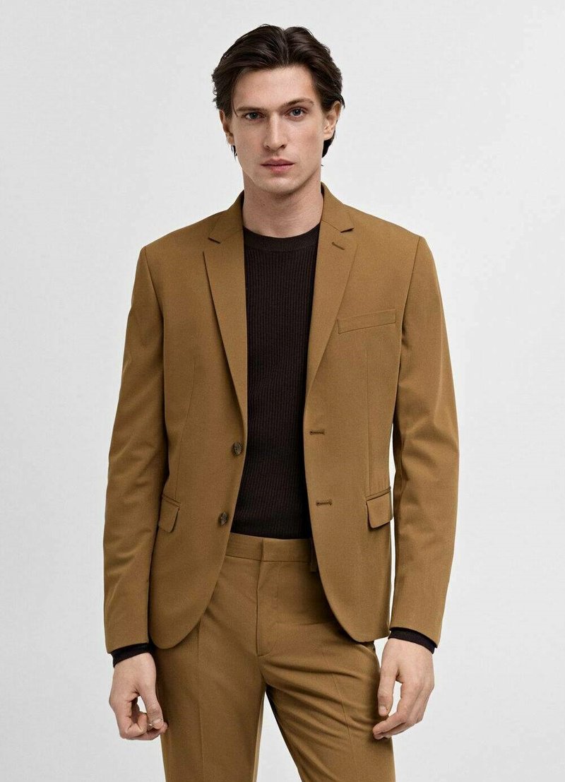 Brown tailored suit with a single-breasted blazer and slim fit trousers, featuring notch lapels and a chest pocket, over a dark ribbed sweater.