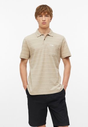 Junger Mann mit Händen in den Taschen, trägt ein beiges kurzärmeliges Poloshirt und schwarze Shorts vor weißem Hintergrund.
