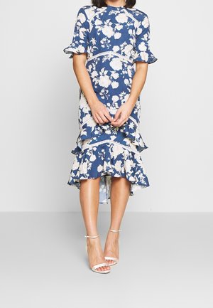 Vestito blu a fiori di lunghezza fino al ginocchio con maniche e orlo arricciati, indossato con sandali bianchi con cinturino alla caviglia e tacco alto, mani giunte davanti.