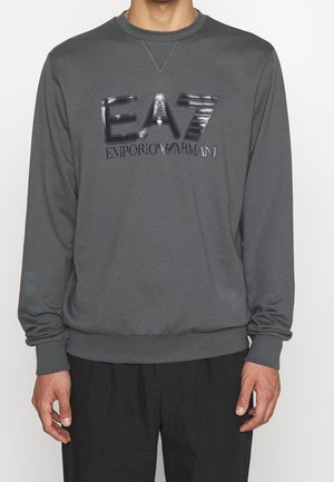 Mężczyzna w szarej bluzie z błyszczącym, czarnym logo "EA7 Emporio Armani" na piersi, w połączeniu z czarnymi spodniami.