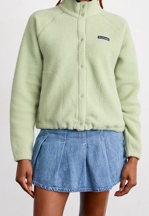 Frau trägt hellgrüne Columbia-Fleecejacke mit Druckknöpfen und blauen plissierten Jeans-Minirock, steht mit entspannten Armen.