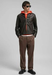 Giacca di pelle nera con chiusura frontale a zip, indossata sopra una felpa con cappuccio arancione. Pantaloni marroni plissettati e scarpe nere completano il look. Occhiali da sole indossati.