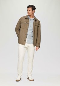 Lichte bruine jas met knoopsluiting, twee voorzakken; gecombineerd met een lichtblauw geruit overhemd, crèmekleurige broek en casual sneakers.