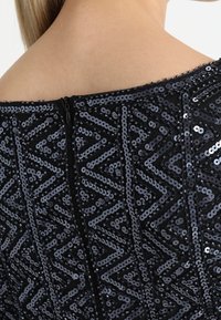 Detailní záběr na záda osoby v černých šatech bez ramínek se vzorem ze zigzag flitrů a centrálním zipem.