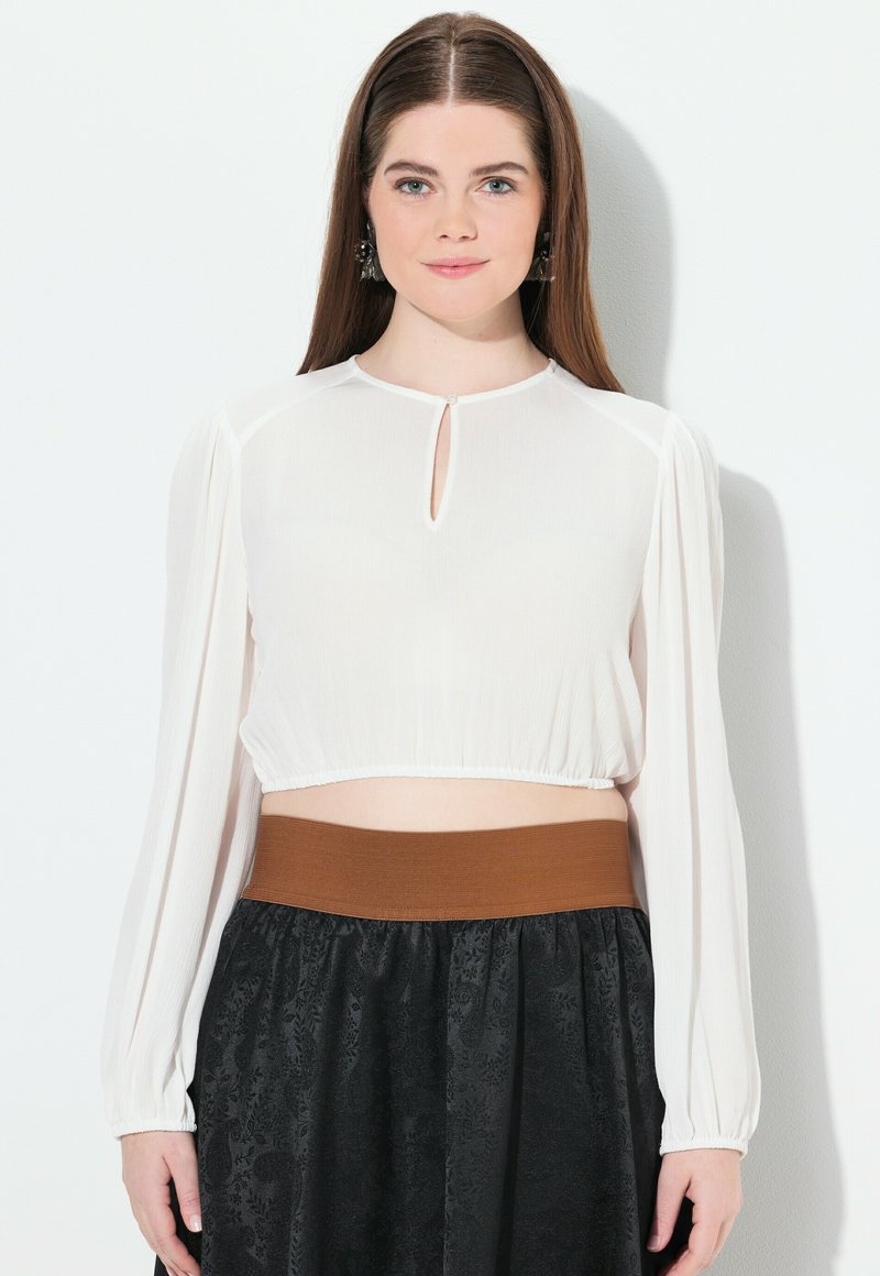 Blusa blanca recortada con mangas largas y escote en forma de llave, combinada con una falda negra texturizada y una cinturilla elástica marrón.