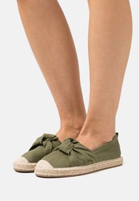 Espadrilles vert olive avec un design en tissu noué, dotées d'une semelle en corde de jute naturel. Texture douce et forme de bout rond.