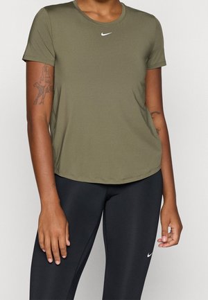 Person trägt ein olivgrünes Nike kurzärmeliges Sportshirt und schwarze Nike-Leggings, steht vor einem einfarbigen Hintergrund.