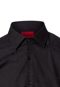 Zwarte shirt met een textuurafwerking, voorzien van een klassieke kraag, knoopsluiting en een rood label met "HUGO" aan de binnenkant van de halslijn.