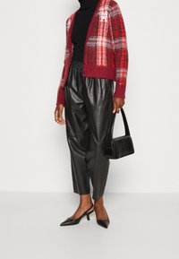 Cardigan à carreaux rouge et blanc avec des manches longues, associé à un pantalon en cuir noir taille haute et des chaussures à talons aiguilles noires. Sac à main noir inclus.