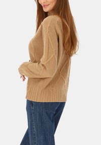 Beżowy sweter z grubym splotem w romby o fakturze, z prążkowanym wykończeniem u dołu i przy mankietach, zestawiony z ciemnoniebieskimi dżinsami, pokazany z boku.