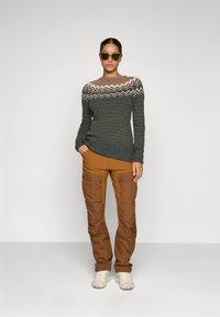 Sweater in dark green with white, brown, and tan zigzag pattern; paired with brown cargo pants and beige footwear.