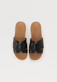Sorte slidesandaler med en teksturert overdel som har utskårne design og en dekorativ stropp. Lysebrune innersåler gir kontrast og komfort.