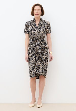 Nainen, jolla on lyhyet ruskeat hiukset, pukeutuneena tummansiniseen mekkoon, jossa on beige kukkakuosi ja lyhyet hihat, sekä kermaisen väriset kengät, seisomassa valkoista seinää vasten.