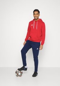 Roter Kapuzenpullover mit weißen Akzenten und Kängurutasche, kombiniert mit marineblauen Jogginghosen mit Logo. Schwarze Sportschuhe. Fußball zu Füßen.