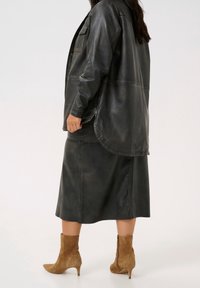 Sort læder oversized skjortejakke parret med en matchende nederdel og brune ankelstøvler. Glat tekstur med synlige sømdetaljer.