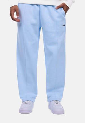 Personne portant un pantalon ample bleu clair avec des poches latérales et des baskets blanches, debout les mains détendues.