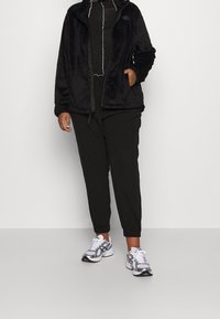 Persona che indossa una giacca in pile nera, pantaloni neri e sneakers bianco-grigie, in piedi su uno sfondo grigio chiaro uniforme.