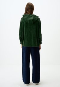 Felpa verde con cappuccio, dal texture morbido, vestibilità ampia e spalle scese, abbinata a pantaloni larghi blu navy e scarpe bianche.