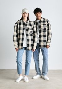 Juoda ir kremiškai dryžuota flanelinė striukė su krūtinės kišenėmis, dėvima ant balto marškinėlių; derinama su šviesiai mėlynomis džinsais ir baltais sportbačiais.