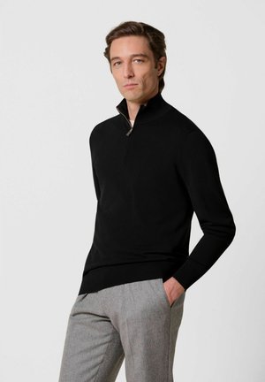 Hombre con cabello corto y castaño que lleva un suéter negro con cremallera y pantalones grises, de pie con una mano en el bolsillo frente a un fondo liso.